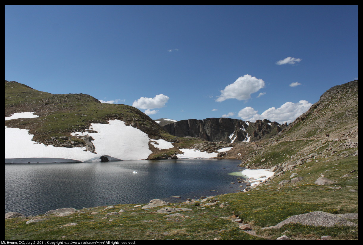 Lake above the tree line on Mt. Evans with a chunk of white snow floating in blue water and white clouds in the blue sky above the mountain.