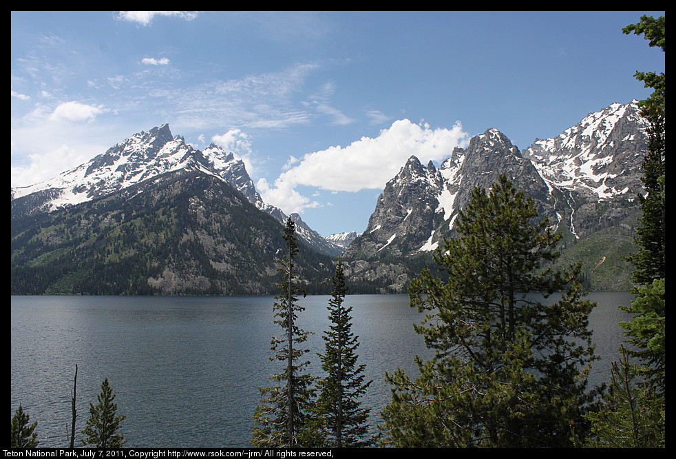 Rocky mountain peaks with snow behind a lake and trees.